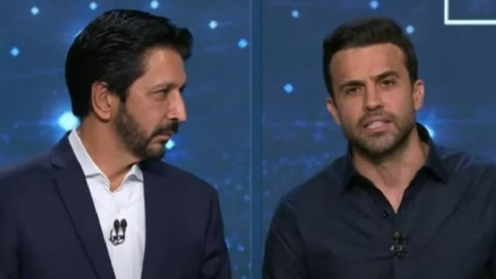 Dois homens em estúdio de TV, um de terno e outro de camisa escura, ambos com microfones