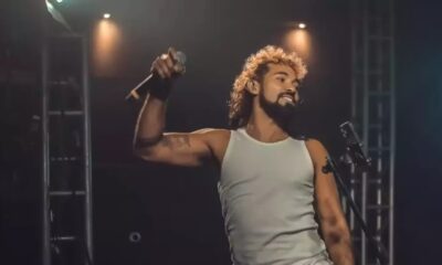 Homem com cabelo encaracolado e barba, em camiseta branca, canta com microfone no palco