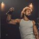 Homem com cabelo encaracolado e barba, em camiseta branca, canta com microfone no palco