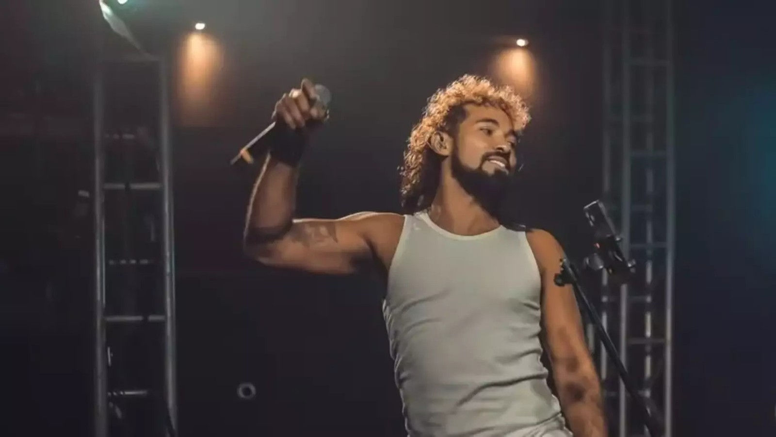 Homem com cabelo encaracolado e barba, em camiseta branca, canta com microfone no palco