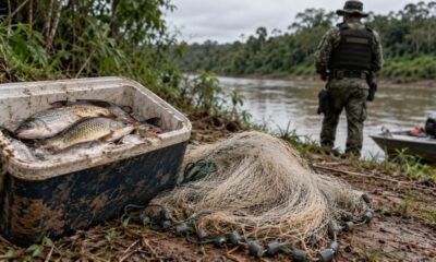 Peixes em cooler, rede de pesca no chão, policial militar e barco à beira de rio