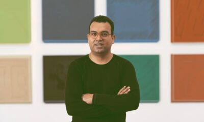 Homem de óculos, camisa preta, braços cruzados, em frente a painéis artísticos coloridos na parede