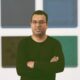 Homem de óculos, camisa preta, braços cruzados, em frente a painéis artísticos coloridos na parede