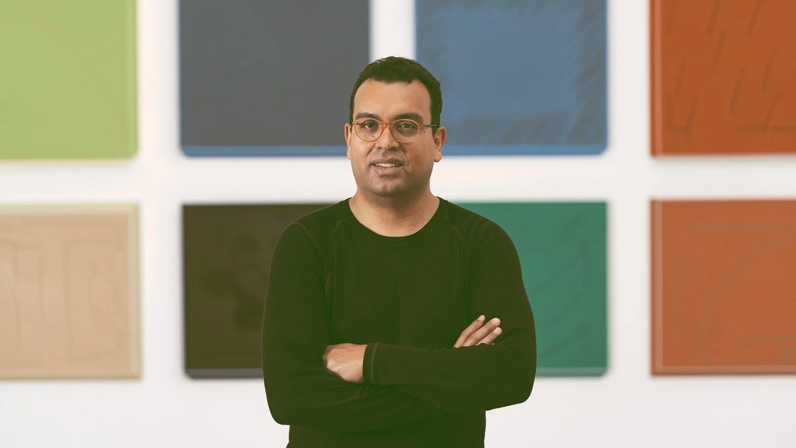 Homem de óculos, camisa preta, braços cruzados, em frente a painéis artísticos coloridos na parede