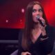 Anitta com cabelo liso, vestindo preto, segurando microfone, fundo vermelho com #EstrelasDaCasa