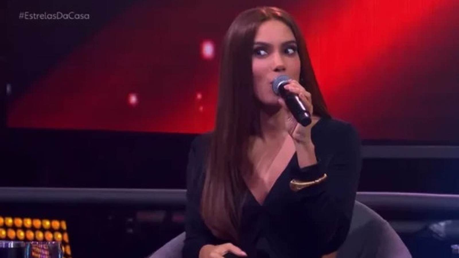 Anitta com cabelo liso, vestindo preto, segurando microfone, fundo vermelho com #EstrelasDaCasa