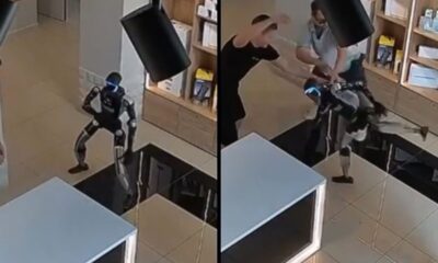 Robô humanoide preto com luz azul na cabeça atacando dois homens em uma loja