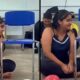 Jovem sentada no chão de sala de aula com expressão emocionada, homem ao fundo e cadeiras azuis