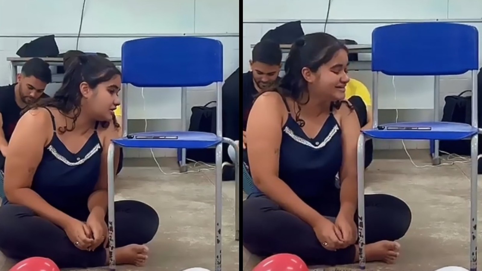 Jovem sentada no chão de sala de aula com expressão emocionada, homem ao fundo e cadeiras azuis