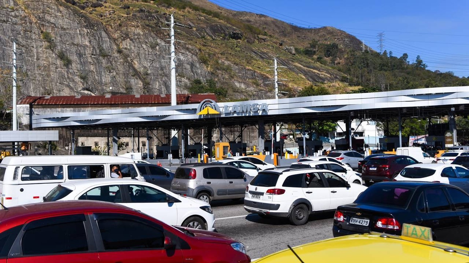 Praça de pedágio LAMSA com múltiplos carros em fila sob estrutura e montanhas