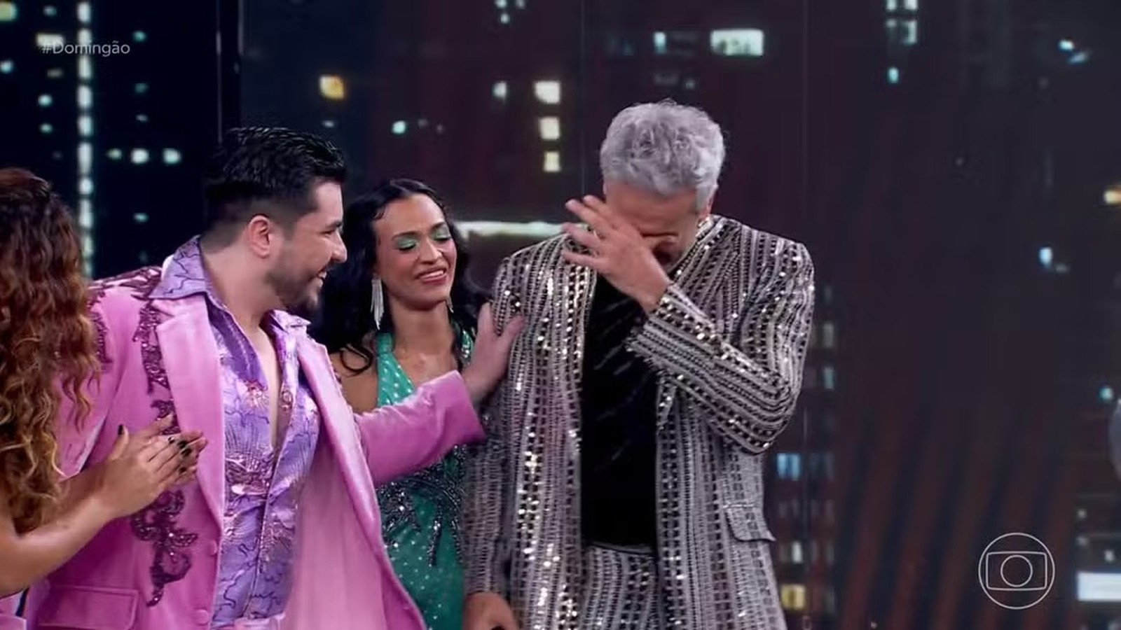 Otaviano Costa, em terno de lantejoulas, cobrindo o rosto emocionado com a equipe no palco