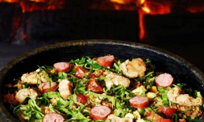 Feijão tropeiro em panela escura com linguiça, torresmo e couve, sobre fundo de fogo
