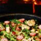 Feijão tropeiro em panela escura com linguiça, torresmo e couve, sobre fundo de fogo