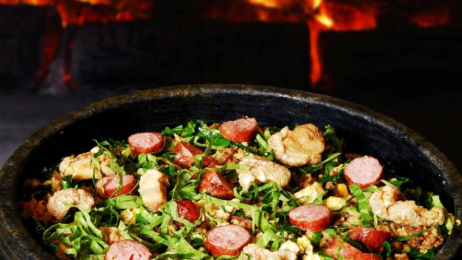 Feijão tropeiro em panela escura com linguiça, torresmo e couve, sobre fundo de fogo