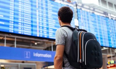 Viajante com mochila de costas olha painel de voos azul em aeroporto; letreiro 'Information'