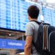 Viajante com mochila de costas olha painel de voos azul em aeroporto; letreiro 'Information'