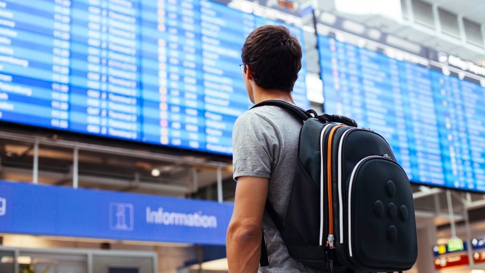 Viajante com mochila de costas olha painel de voos azul em aeroporto; letreiro 'Information'