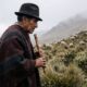 Homem idoso, chapéu, poncho, toca flauta de madeira, ovelhas na montanha