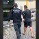 Agente da Polícia Civil escolta homem algemado, vestindo camiseta e shorts escuros