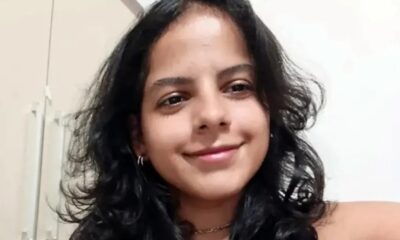 Mulher jovem com cabelo escuro e encaracolado sorri levemente, usando brincos de argola e colar