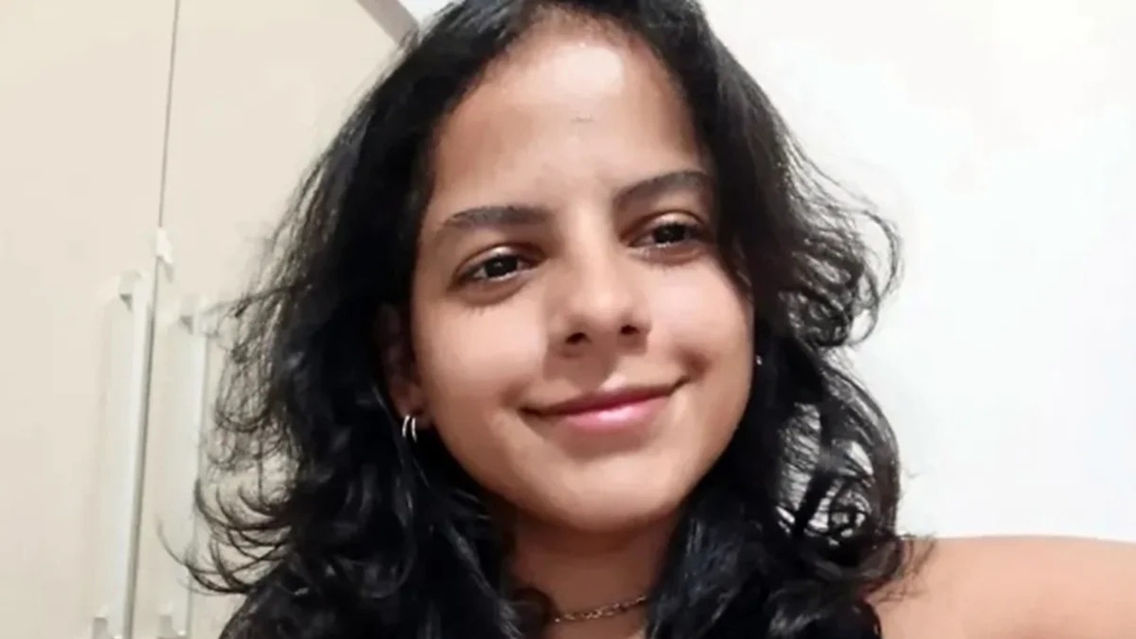 Mulher jovem com cabelo escuro e encaracolado sorri levemente, usando brincos de argola e colar
