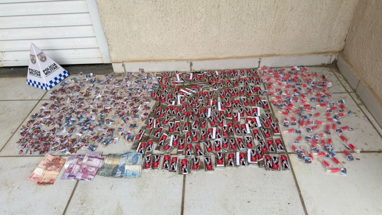 Drogas em embalagens diversas, notas de dinheiro e cone da Polícia Militar em piso claro