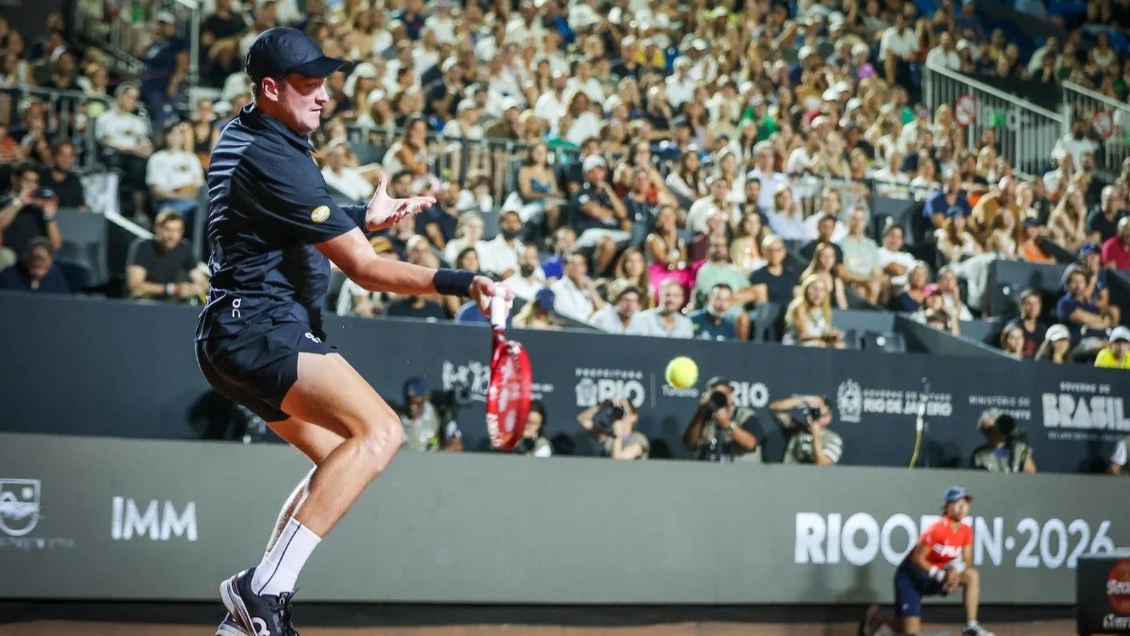 João Fonseca em quadra de saibro com uniforme preto rebatendo bola no Rio Open