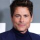 Close-up do ator Rob Lowe, com cabelo castanho, olhos azuis e barba rala, vestindo terno azul