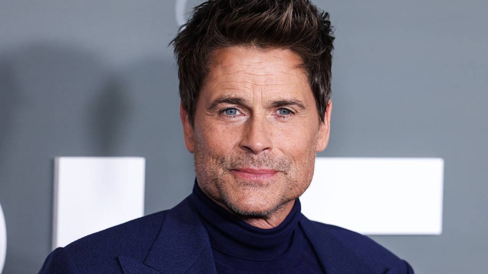 Close-up do ator Rob Lowe, com cabelo castanho, olhos azuis e barba rala, vestindo terno azul