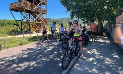 Grupo de ciclistas em ciclovia, ao lado de estrutura de madeira, sob sol forte