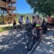 Grupo de ciclistas em ciclovia, ao lado de estrutura de madeira, sob sol forte