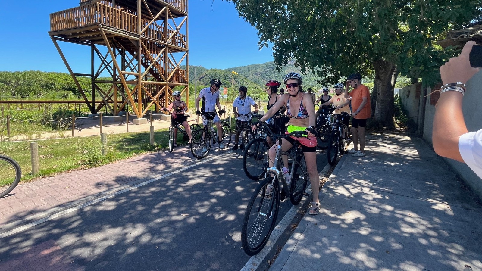 Grupo de ciclistas em ciclovia, ao lado de estrutura de madeira, sob sol forte