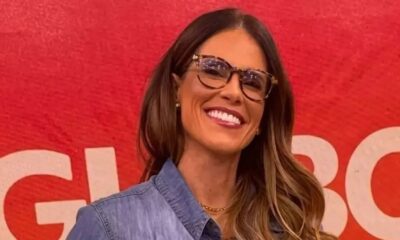 Close-up de Joanna de Assis sorrindo, óculos tartaruga, camisa jeans, fundo vermelho com 'GLOBO'