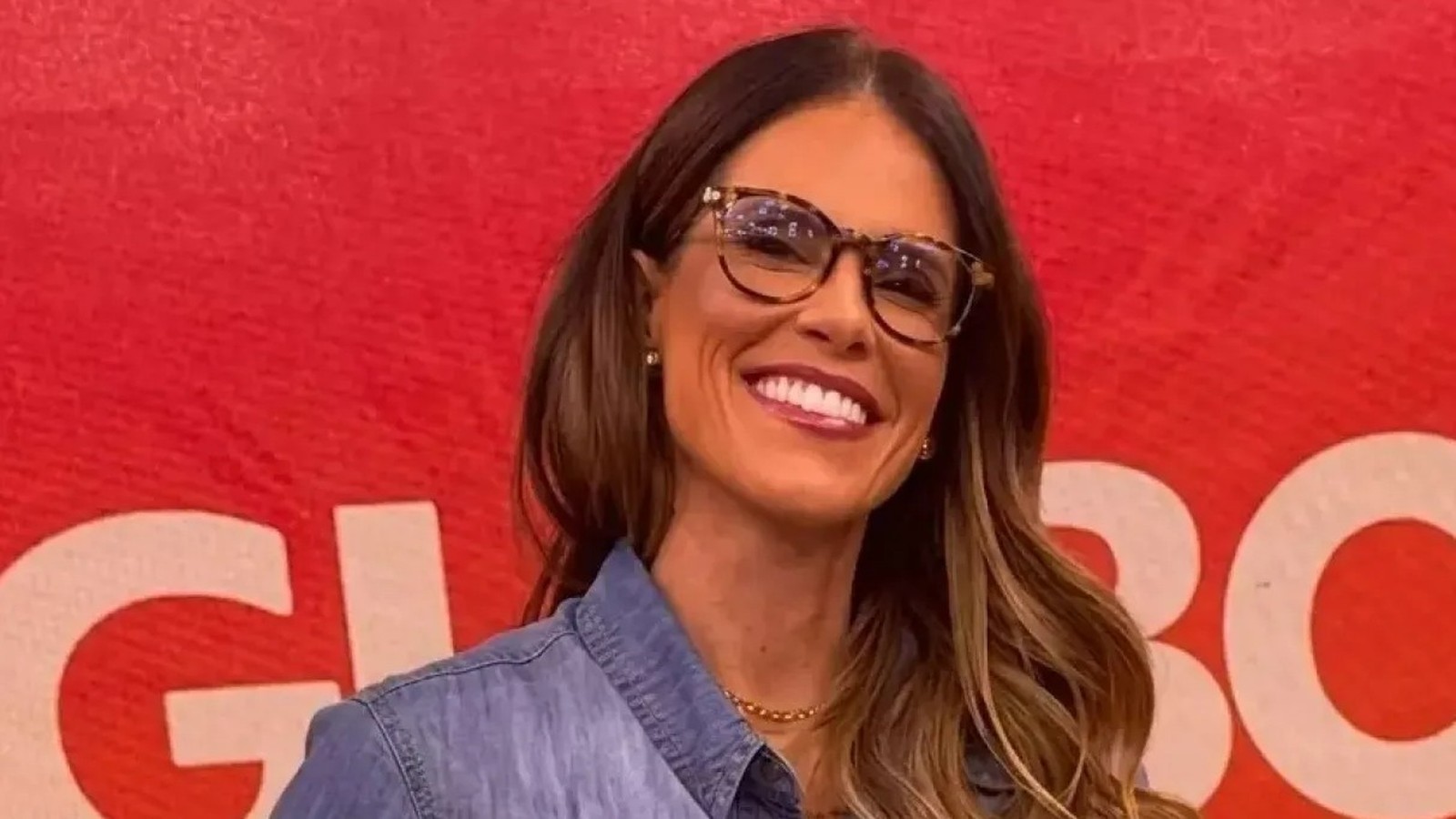 Close-up de Joanna de Assis sorrindo, óculos tartaruga, camisa jeans, fundo vermelho com 'GLOBO'