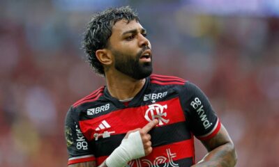 Jogador barbudo com cabelo crespo, camisa vermelha e preta, apontando para o escudo do clube