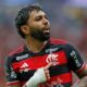 Jogador barbudo com cabelo crespo, camisa vermelha e preta, apontando para o escudo do clube