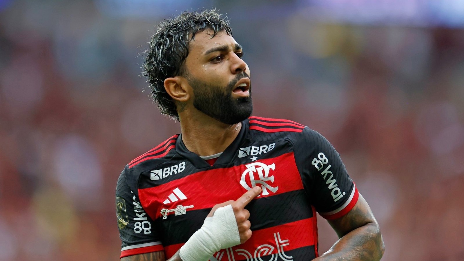 Jogador barbudo com cabelo crespo, camisa vermelha e preta, apontando para o escudo do clube