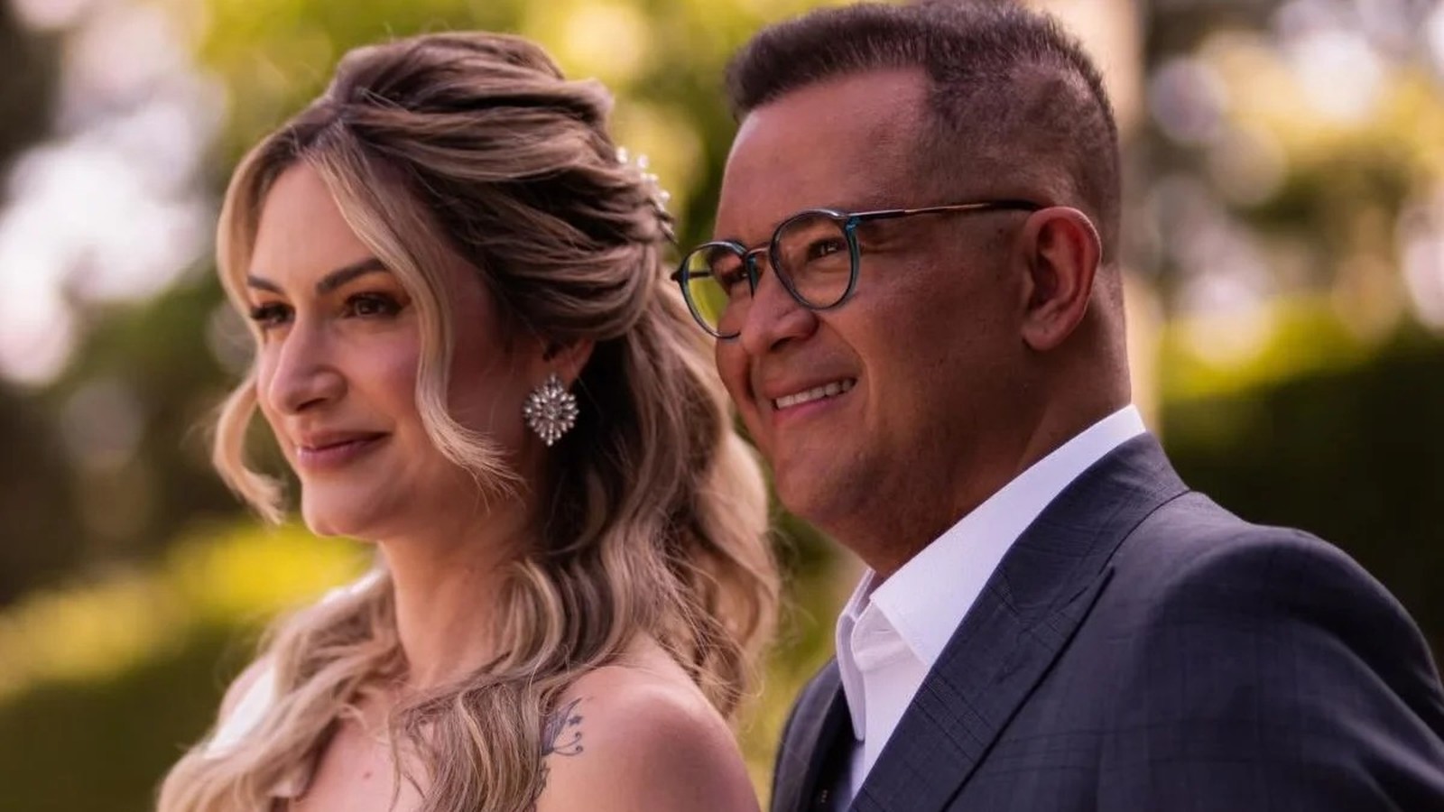 Casal sorrindo em close-up, homem de óculos e terno, mulher com penteado elaborado