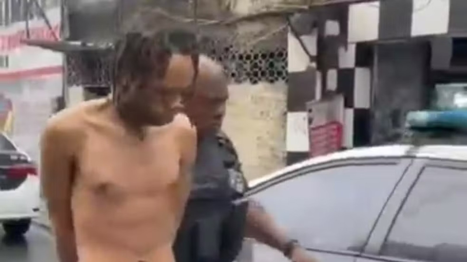 Homem de torso nu com dreadlocks escoltado por policial perto de viatura policial em rua