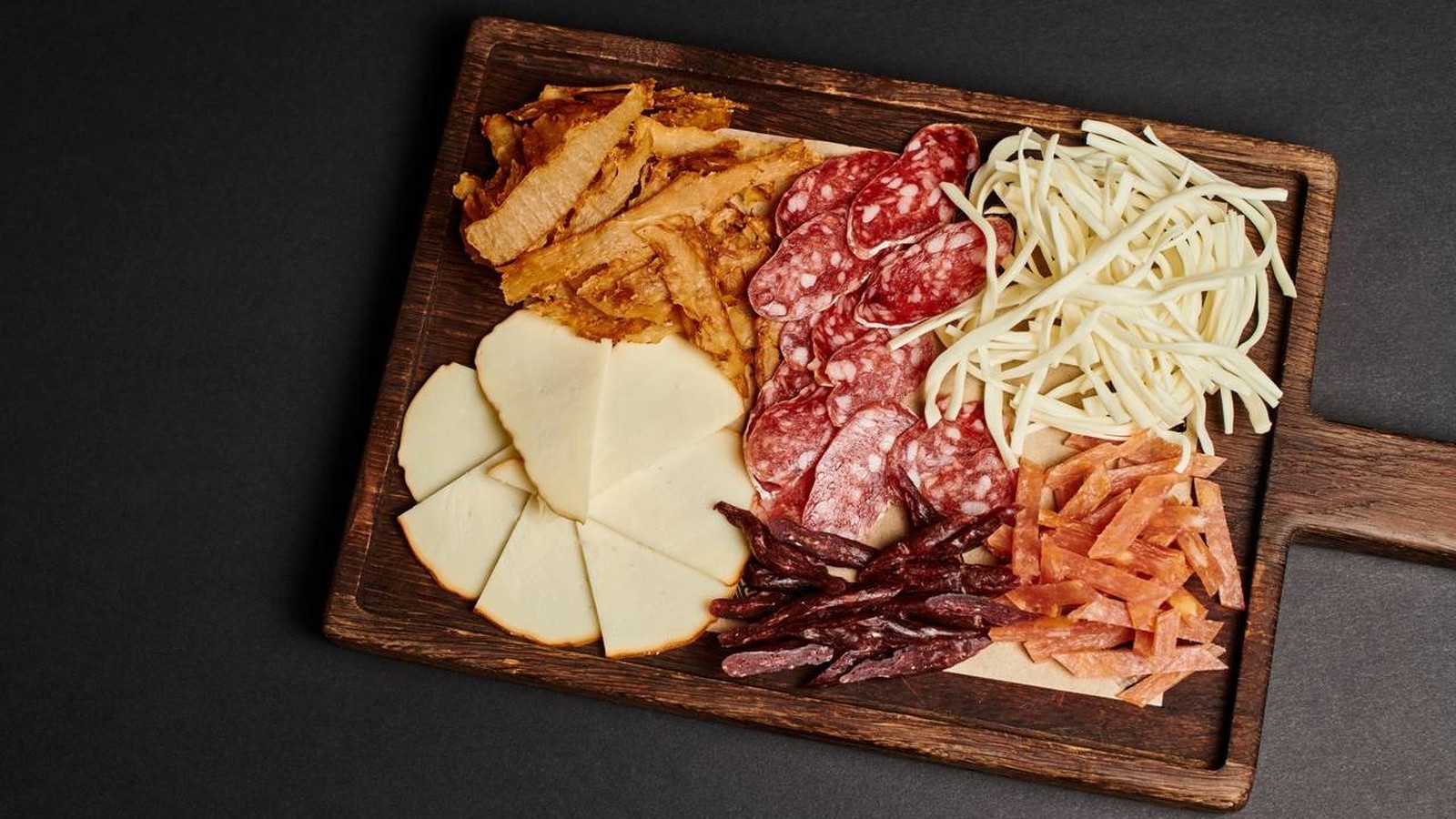 Tábua de madeira com queijos fatiados, queijo desfiado, salame e diferentes tipos de carnes curadas