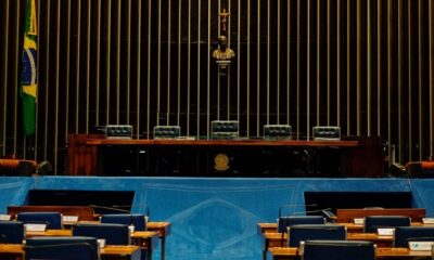 Plenário vazio do Senado Federal com mesas, cadeiras, bandeira brasileira, busto e crucifixo