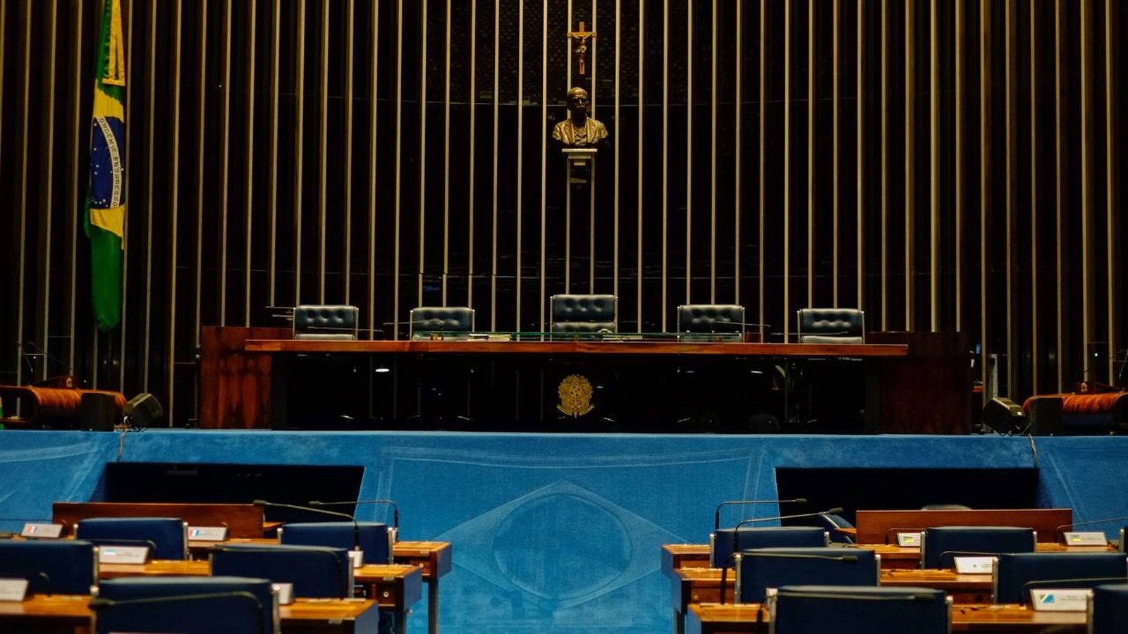 Plenário vazio do Senado Federal com mesas, cadeiras, bandeira brasileira, busto e crucifixo