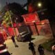 Caminhão de bombeiros com luzes vermelhas em rua noturna, pessoas e casa com brilho vermelho