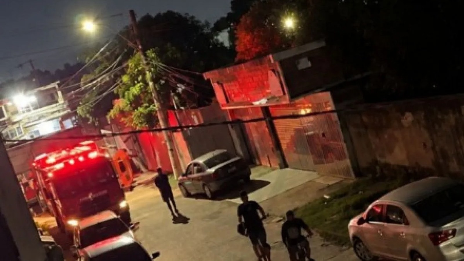 Caminhão de bombeiros com luzes vermelhas em rua noturna, pessoas e casa com brilho vermelho