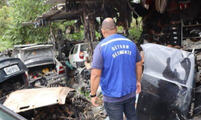 Homem de colete azul "DETRAN DESMONTE" em ferro-velho com carros e peças de automóveis