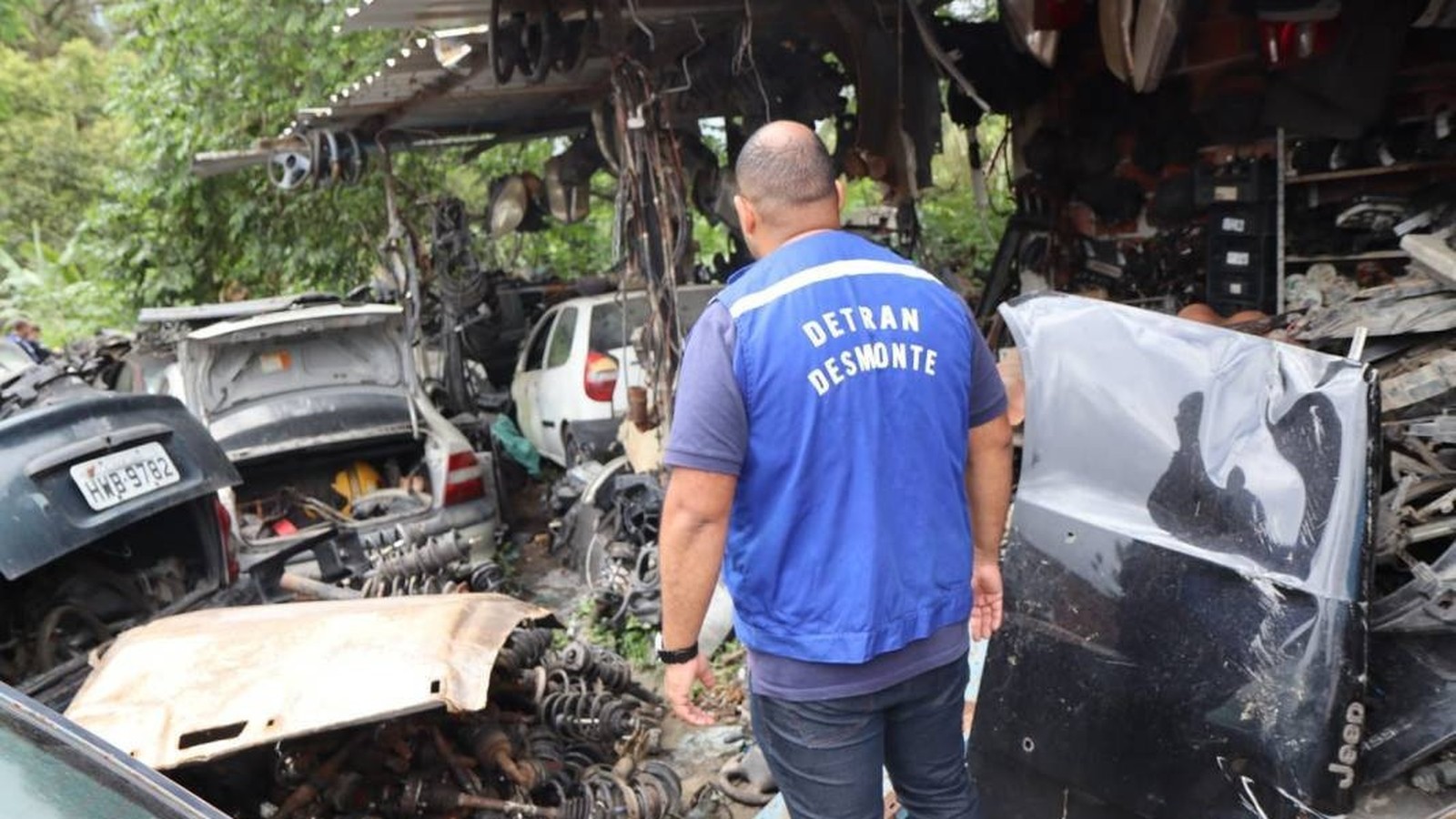 Homem de colete azul "DETRAN DESMONTE" em ferro-velho com carros e peças de automóveis