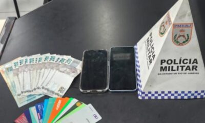 Dinheiro, diversos cartões bancários, dois celulares e placa da PMERJ sobre mesa escura