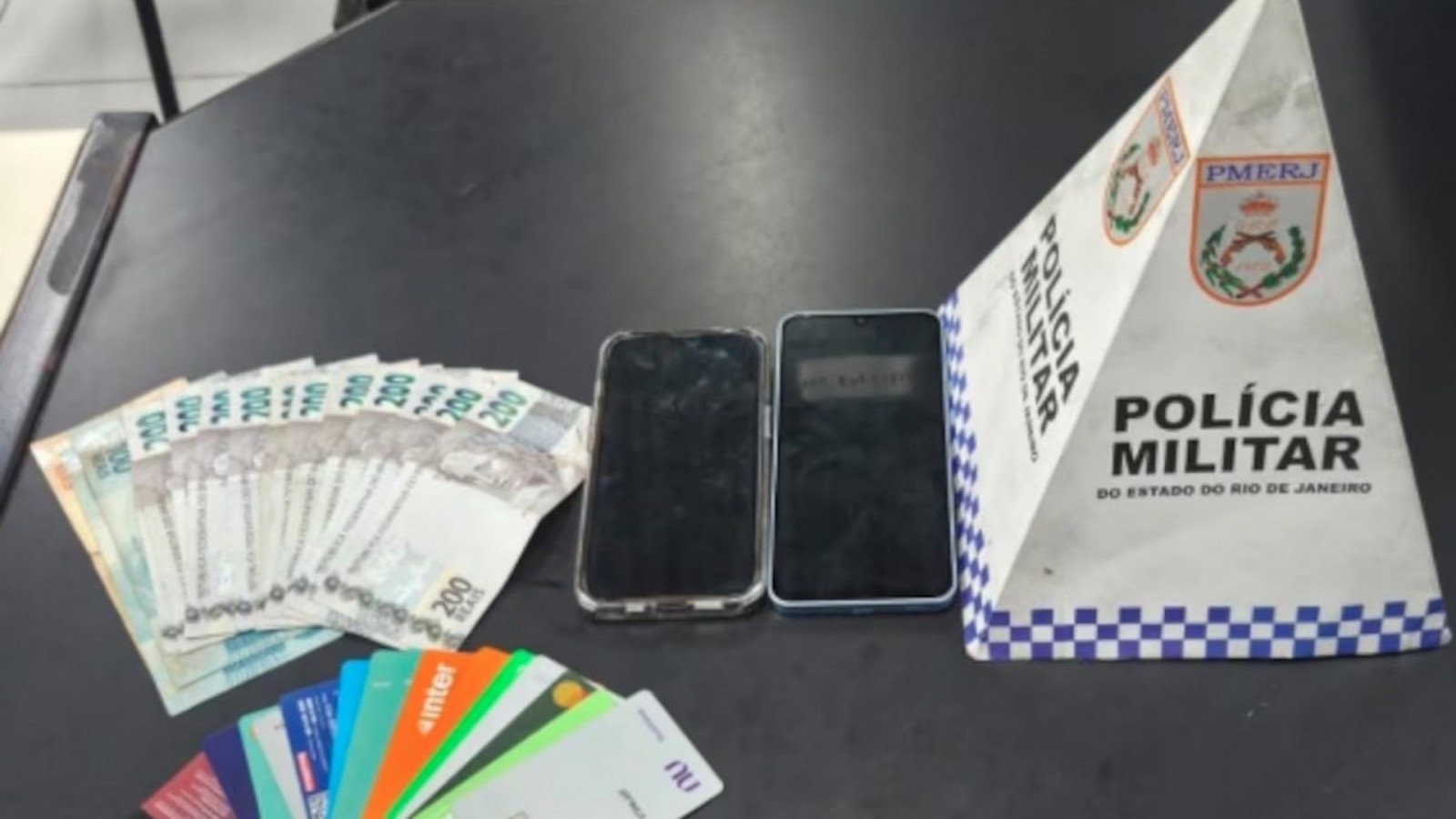 Dinheiro, diversos cartões bancários, dois celulares e placa da PMERJ sobre mesa escura