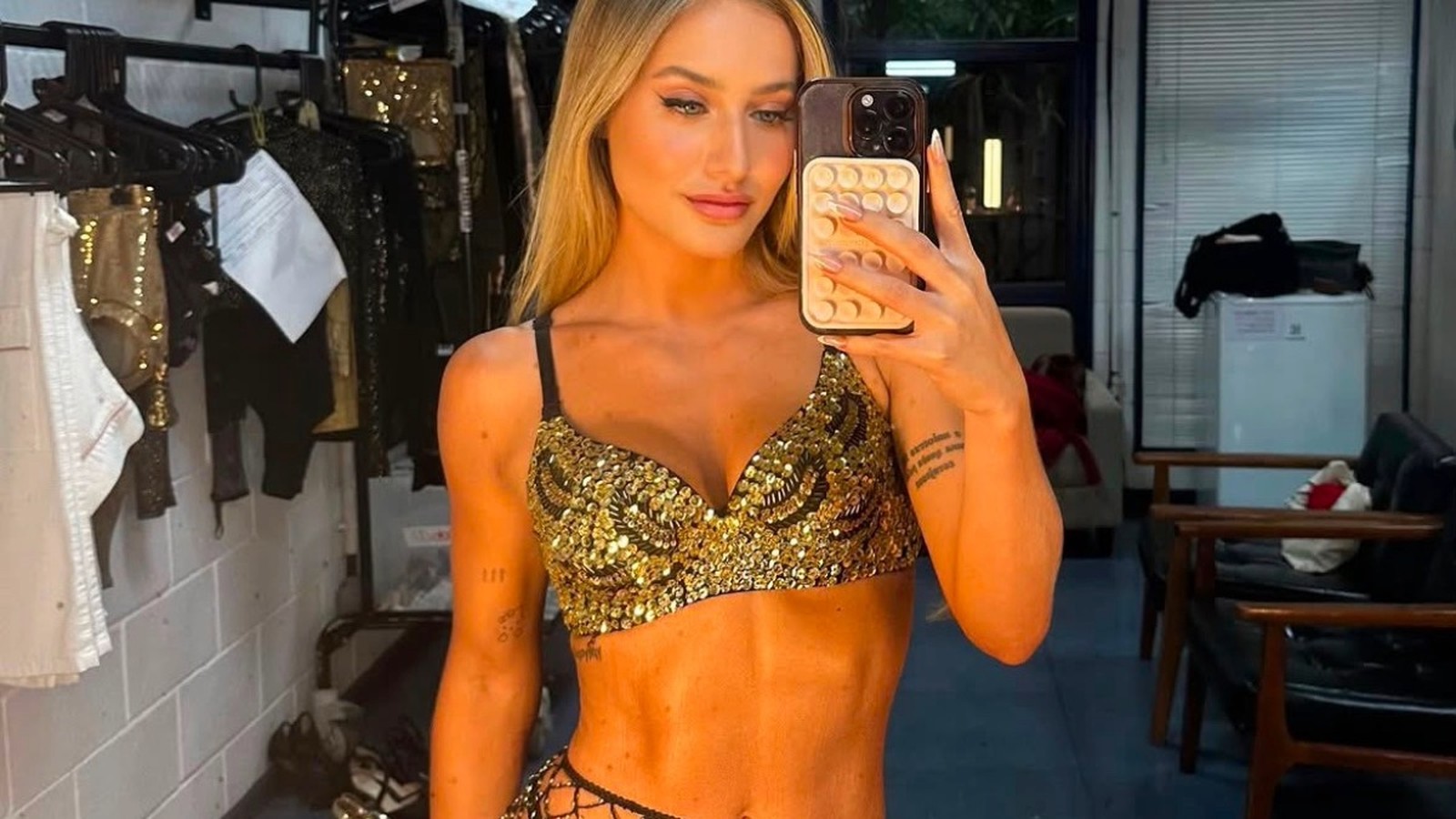 Bruna Griphao em selfie de espelho, vestindo top dourado de paetês, segurando celular, em bastidores