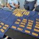 Mesa com pano azul, mostrando barras e pilhas de moedas de ouro, com oficiais